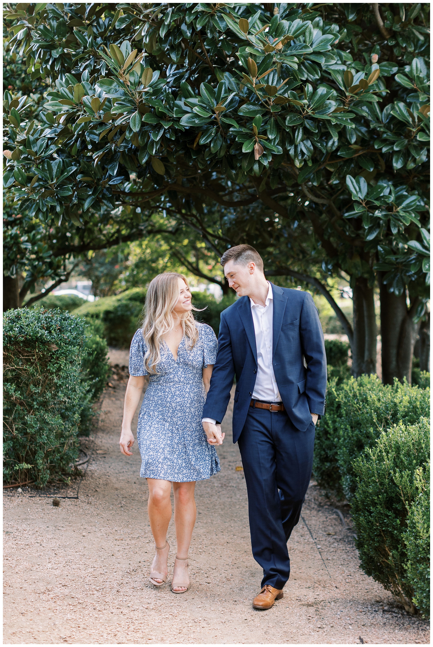 couple in blue dress and blue suit holding hands and walking under a tree in Houston Texas