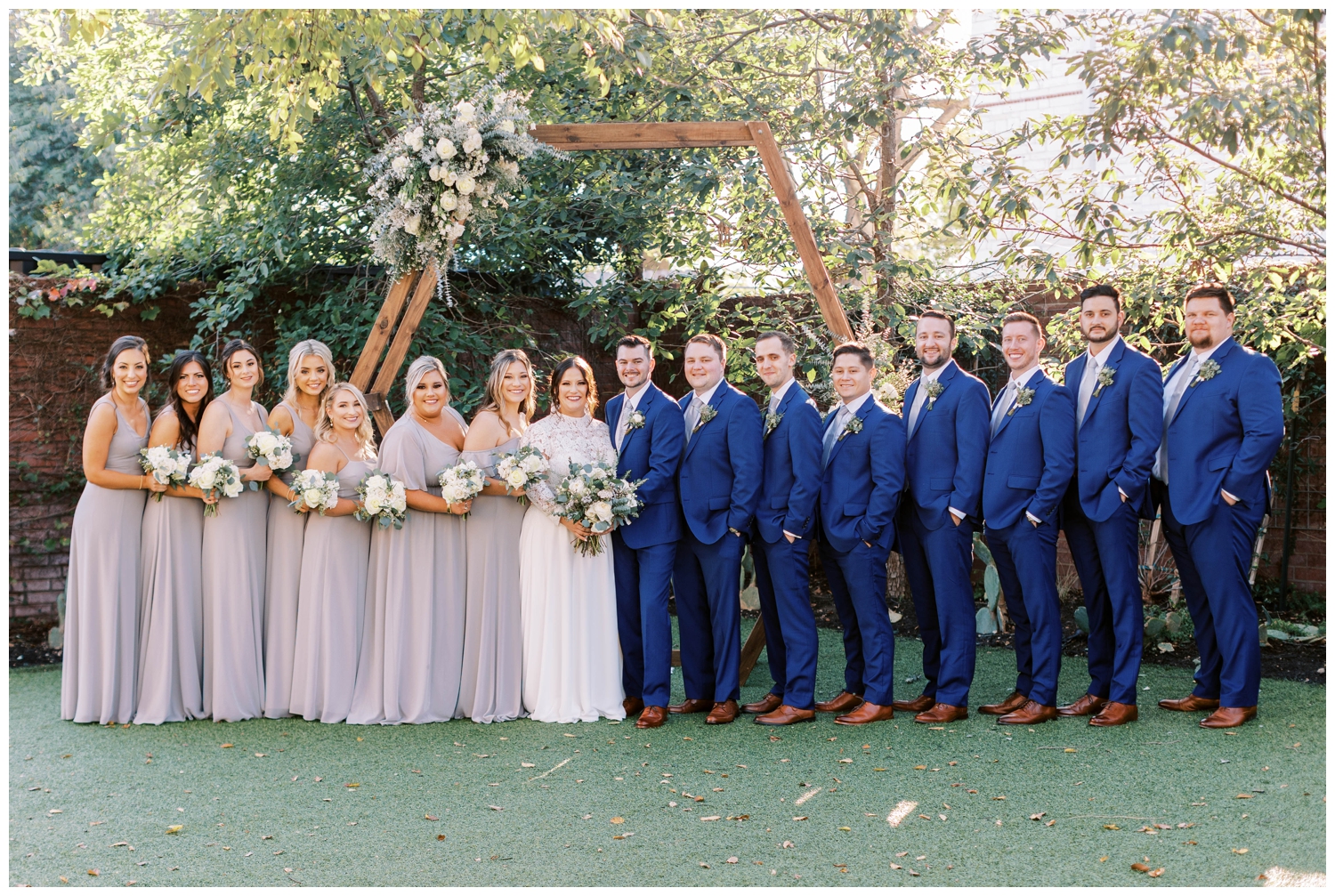 full bridal party portrait standing under hexagon floral arch groom in blue tux portrait standing under tree for Austin outdoor wedding