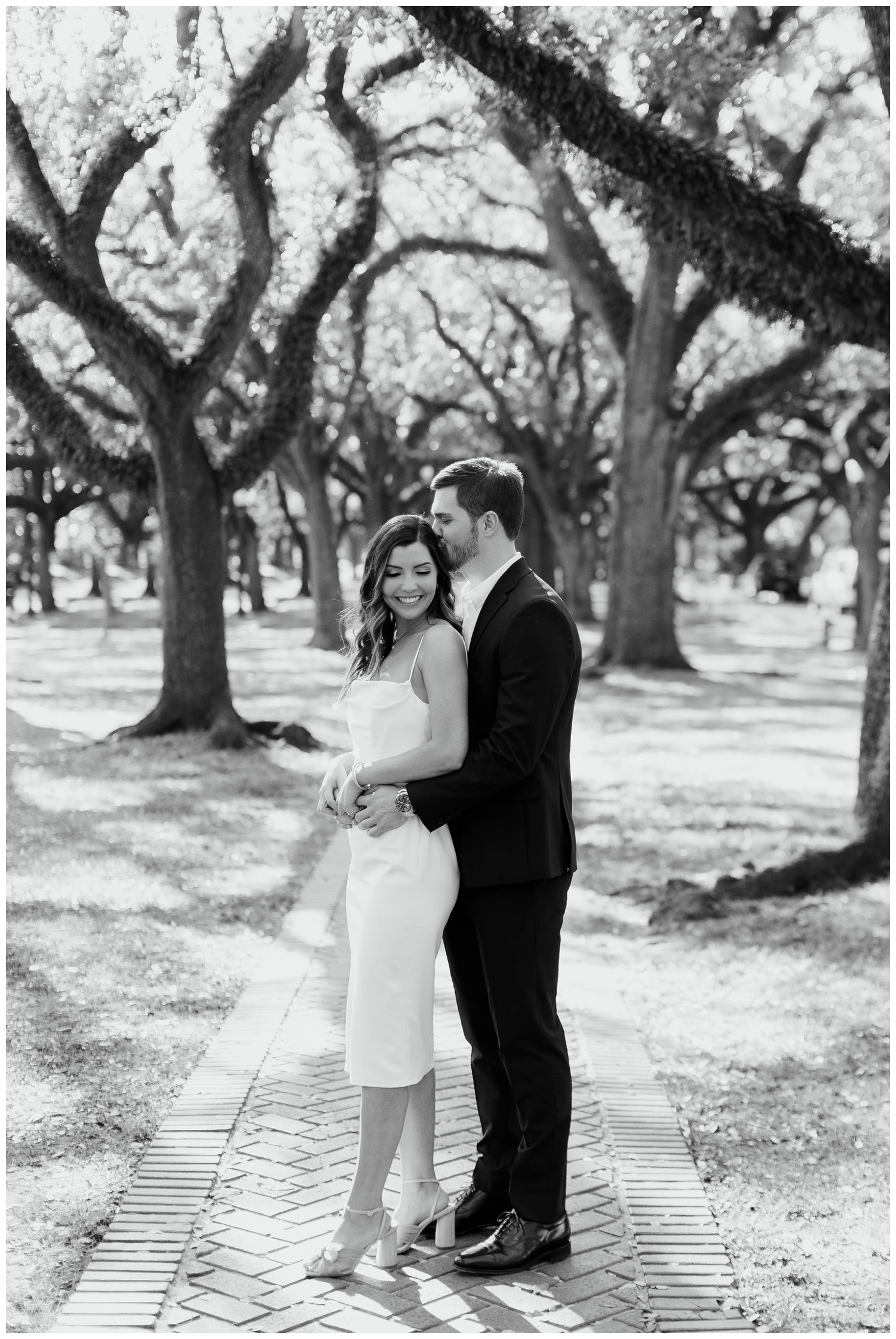 black and white image of engaged couple embracing by tree line on sidewalk