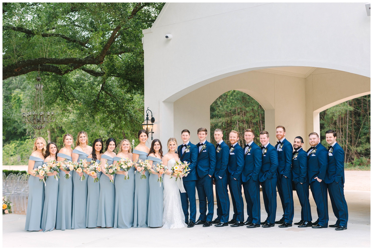 full wedding party standing in a line in blue dresses and blue tuxes outside The Peach Orchard