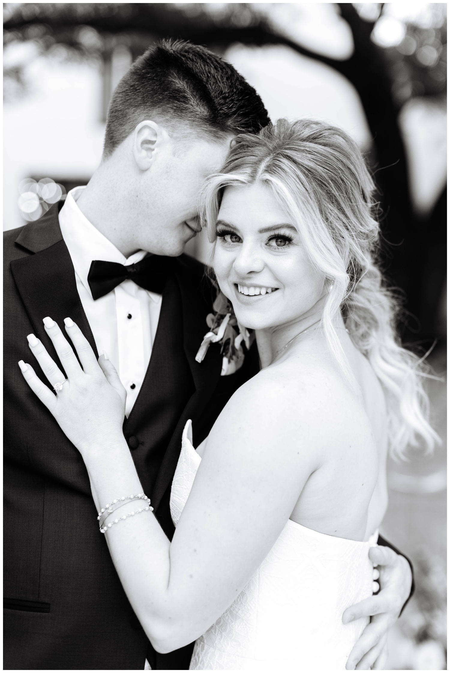 black and white image of bride smiling at camera a groom whispering in her ear