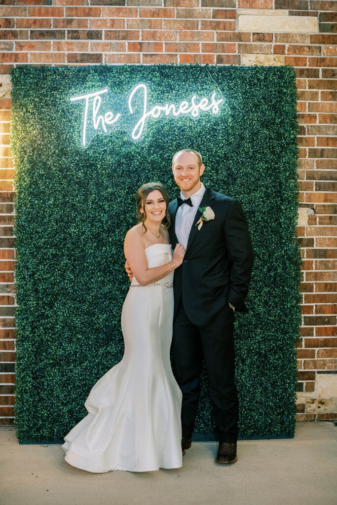 Bride and groom posing in front of greenery with custom name sign