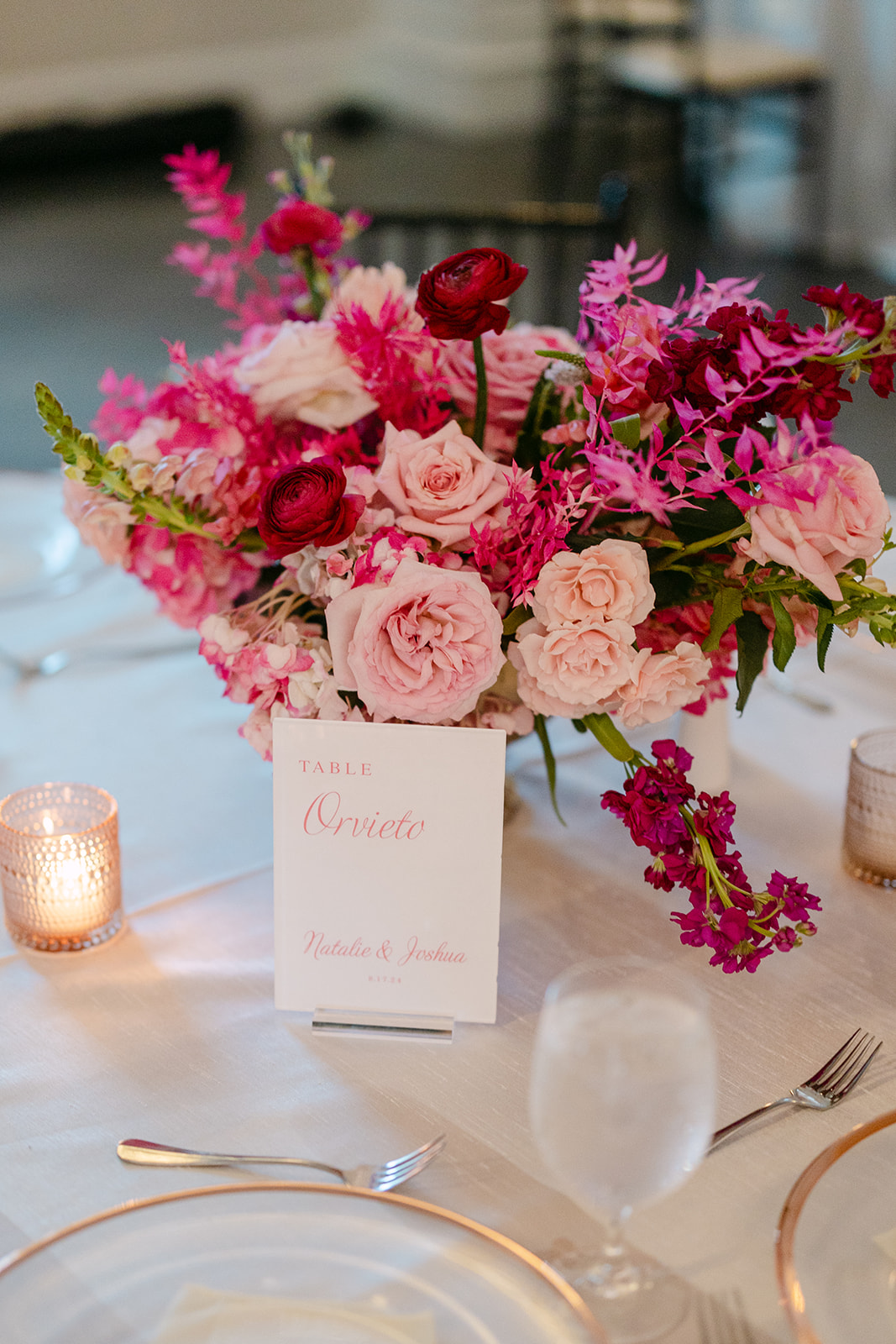 Wedding reception with elegantly set tables featuring tall gold stands with vibrant pink floral arrangements. Black chairs surround the tables, and candles create a soft ambiance.