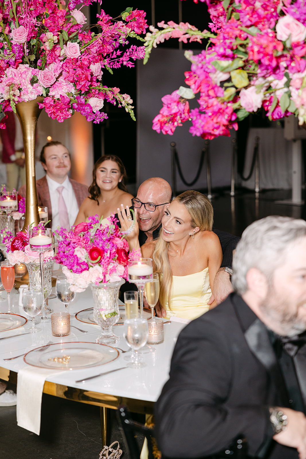 Guests seated at an elegant table decorated with tall arrangements of pink flowers, enjoying drinks and conversation at an event.