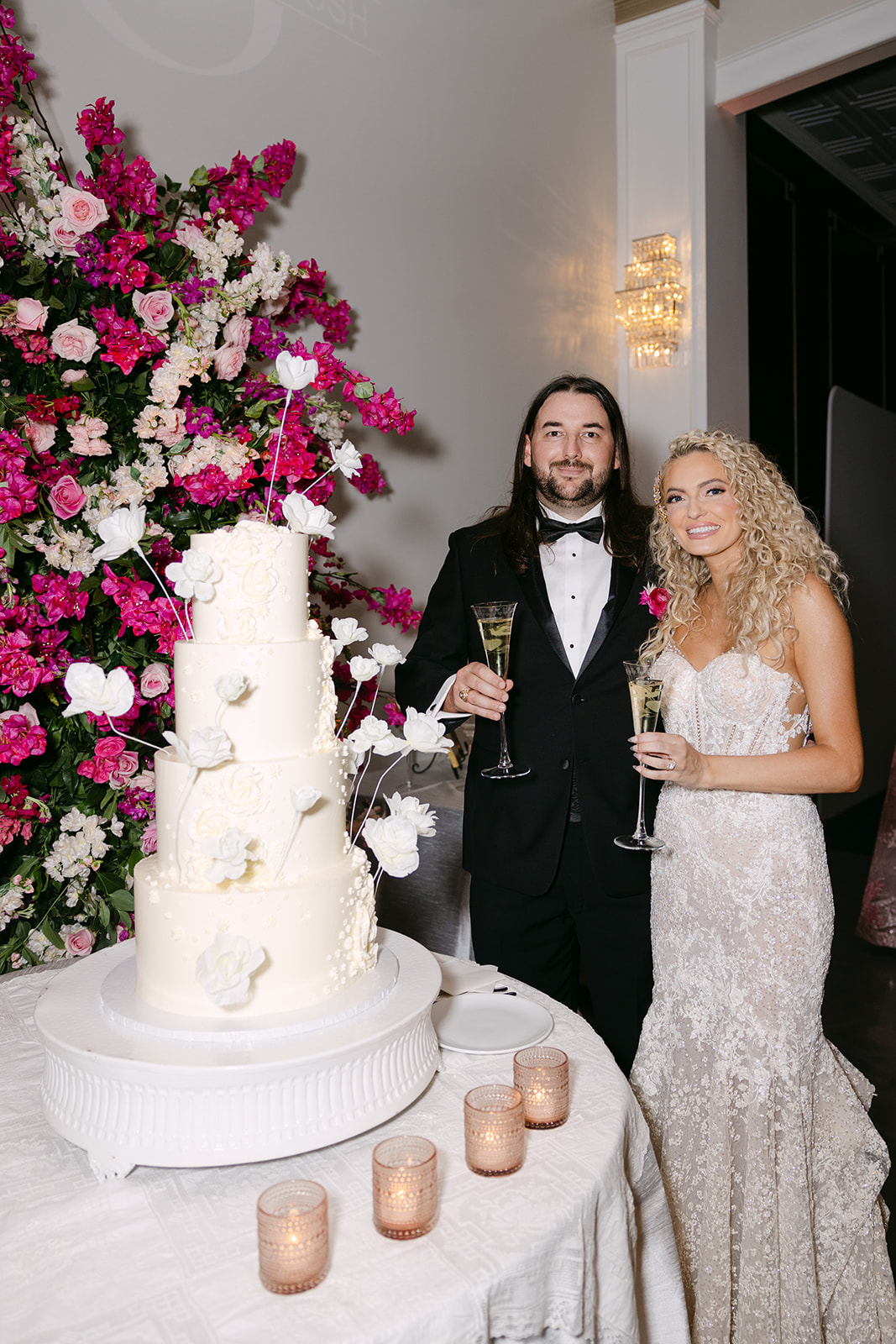 A couple in formal attire stands by a white, tiered wedding cake with floral decorations, holding champagne glasses. A large floral arrangement is in the background.
