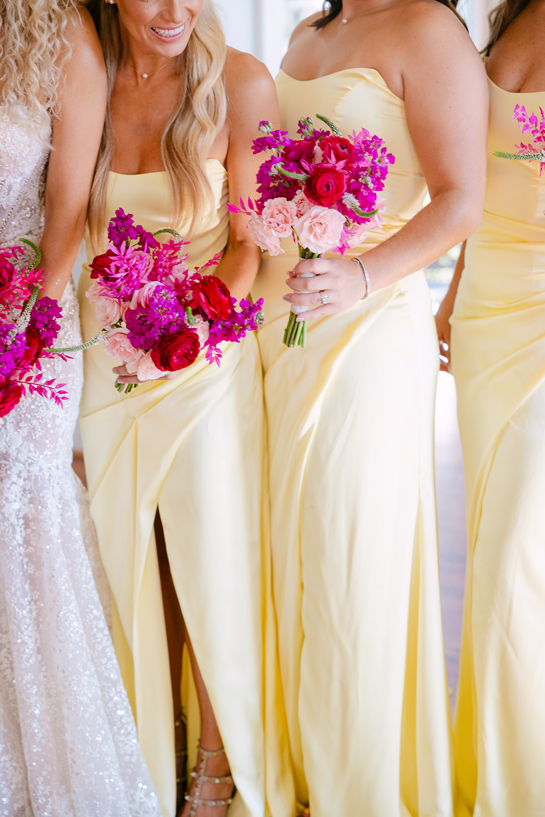A bride in a strapless gown stands with bridesmaids in yellow dresses, all holding pink bouquets, in front of floral arrangements by a window.