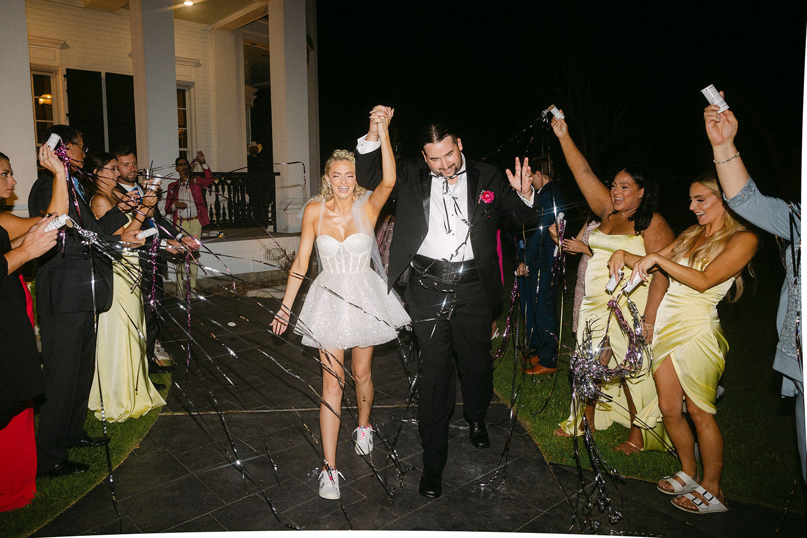 A newlywed couple walks hand in hand outside at night, surrounded by cheering guests tossing ribbons.