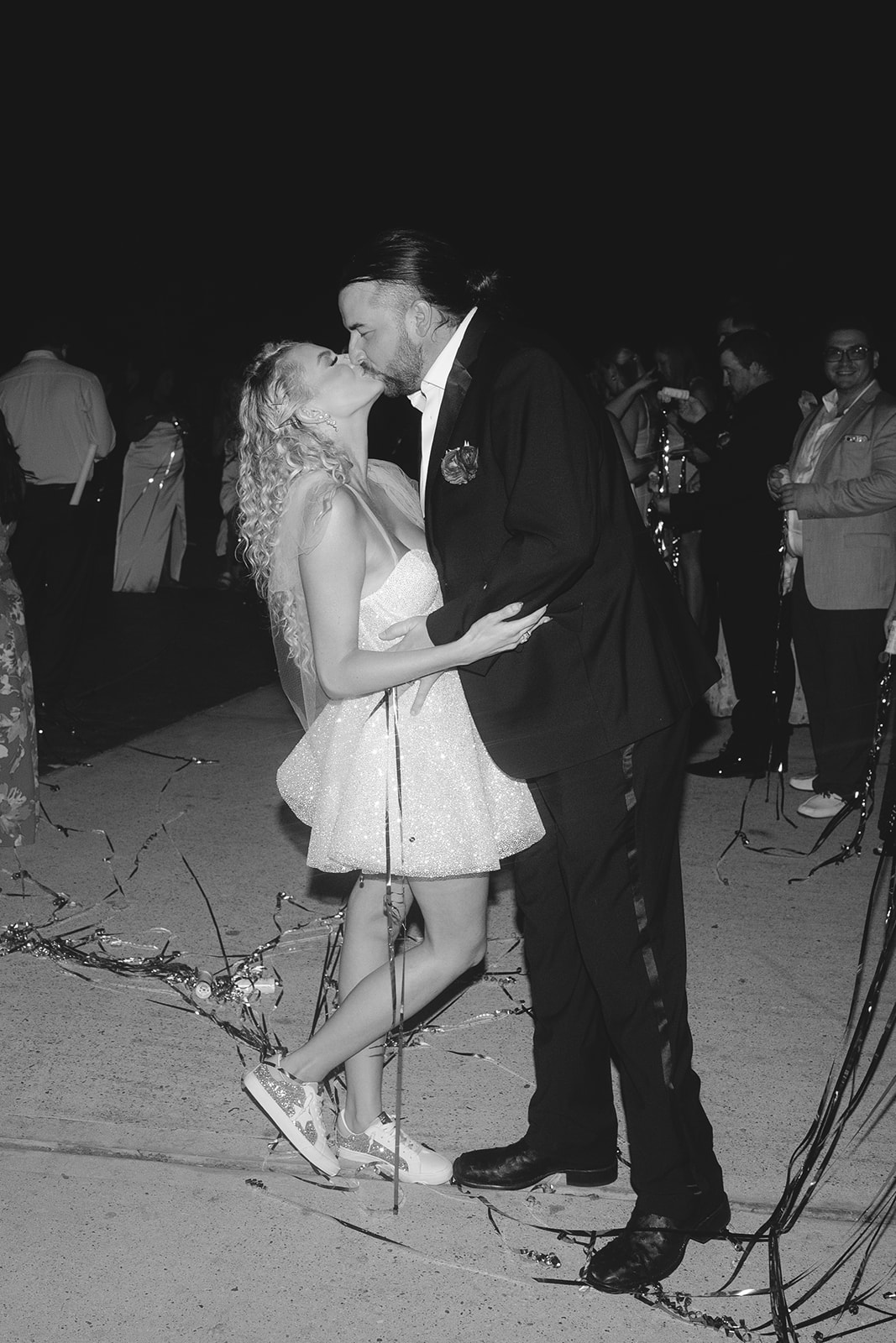 A newlywed couple walks hand in hand outside at night, surrounded by cheering guests tossing ribbons.