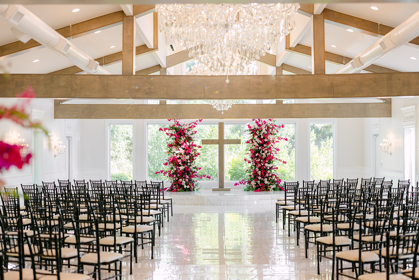 Bright wedding chapel with rows of chairs facing a cross adorned with pink flowers, large windows, and chandeliers.