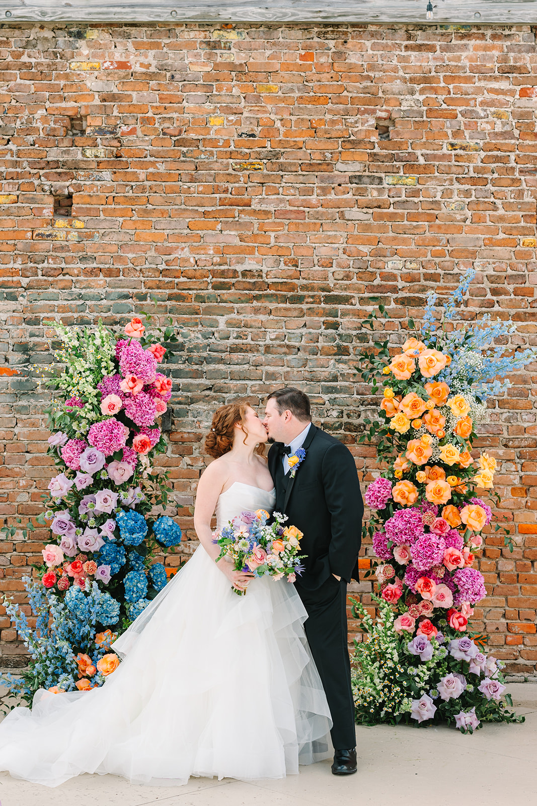 Bride and groom stand smiling in front of a brick wall, surrounded by colorful floral arrangements for their wedding at the artisan in Galveston