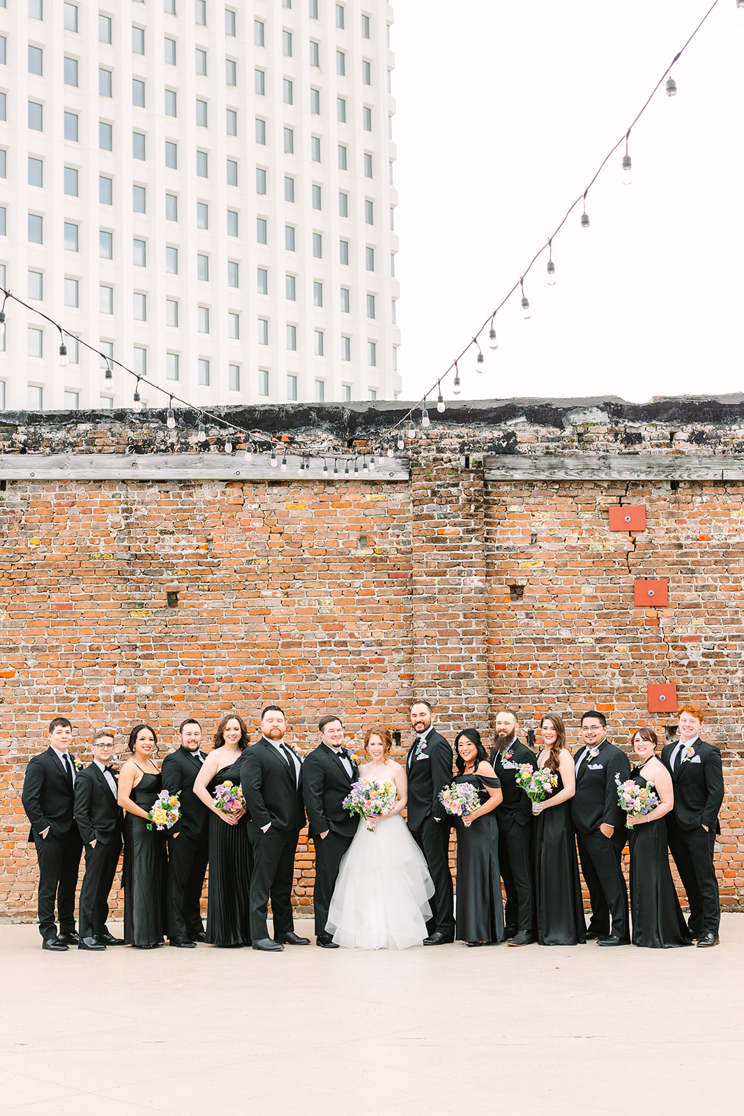 A bride and groom stand close together, surrounded by their wedding party dressed in black attire, holding colorful bouquets, against a brick wall backdrop outdoors for their wedding at the artisan galveston