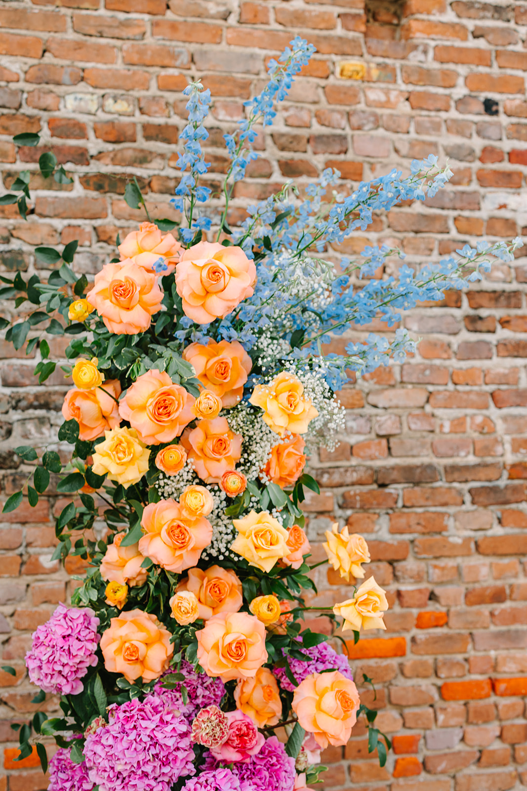 Rows of white chairs face a brick wall decorated with colorful flower arrangements for an outdoor ceremony.