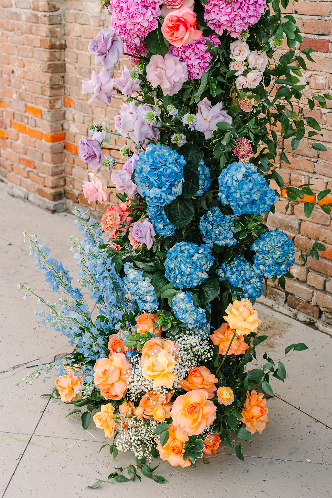 Rows of white chairs face a brick wall decorated with colorful flower arrangements for an outdoor ceremony.