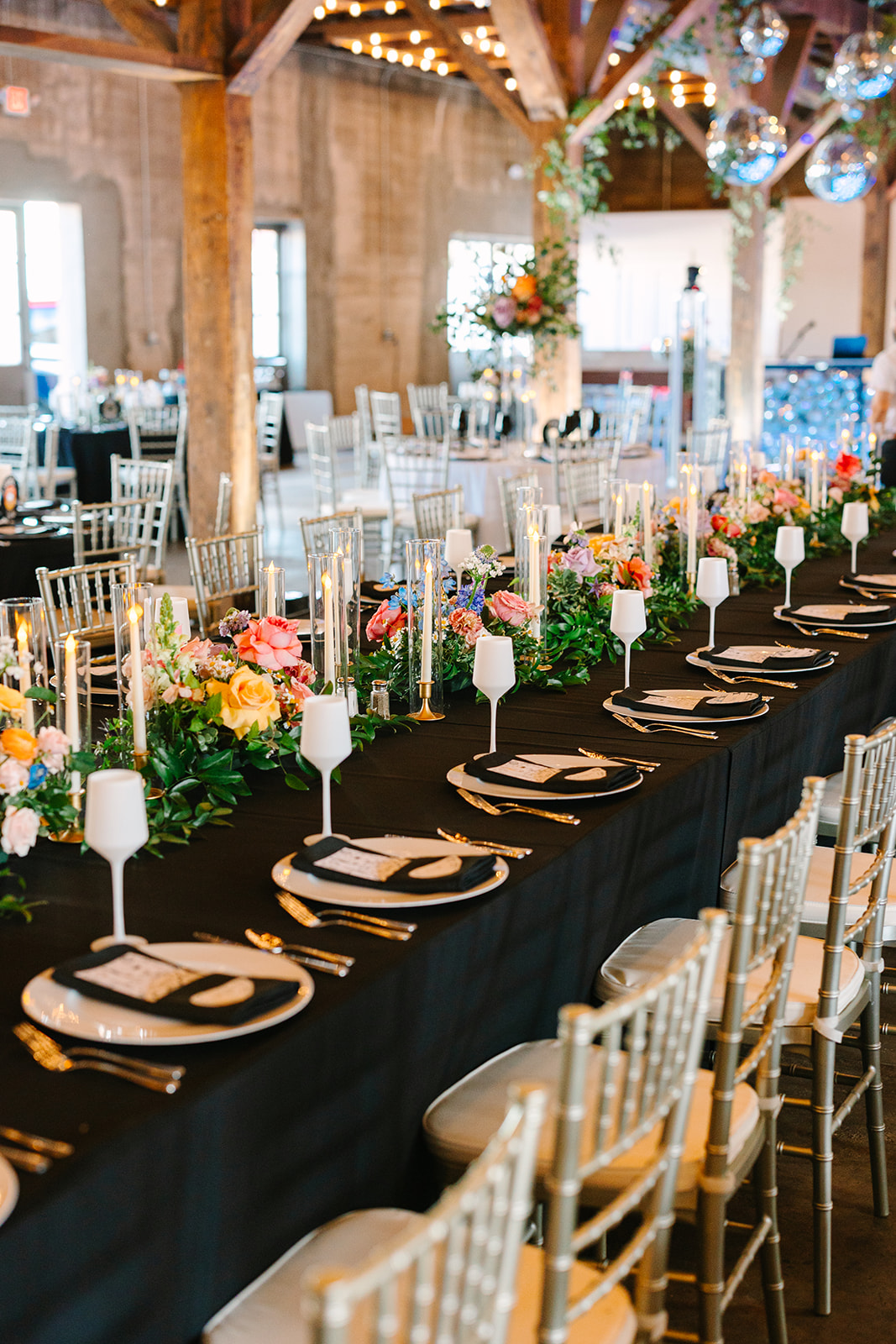 A long banquet table with black tablecloth, white plates, glassware, candles, and colorful floral centerpieces is set for an event in a decorated indoor venue with string lights.