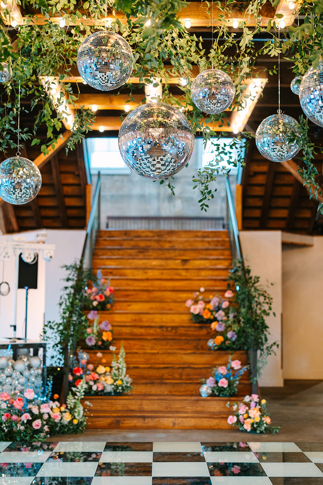 Wooden stairs decorated with colorful flowers and greenery, disco balls hanging from the ceiling, and a checkered black-and-white dance floor below.