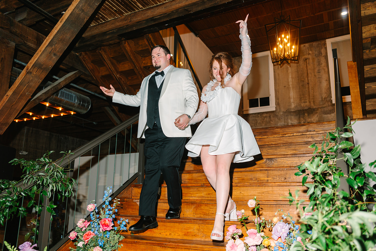 A large group of people dressed formally dance on a colorful light-up floor under disco balls and greenery at a wedding