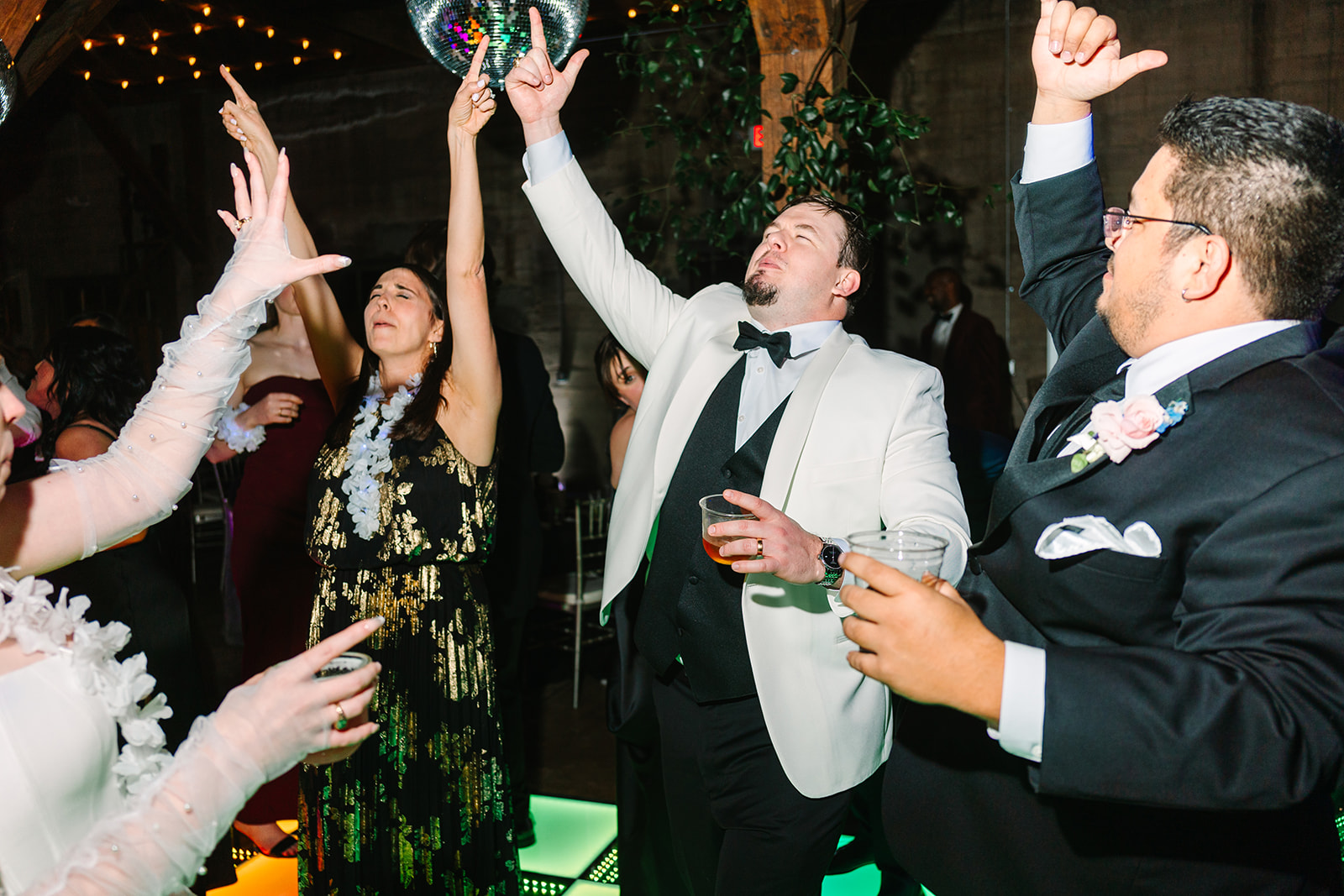A large group of people dressed formally dance on a colorful light-up floor under disco balls and greenery at a wedding