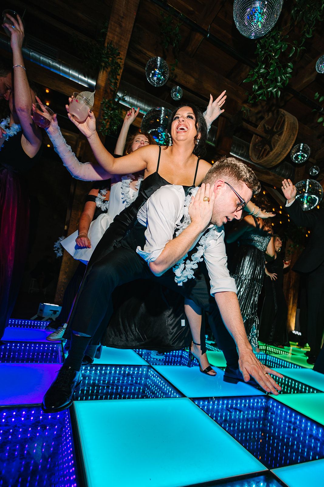 A large group of people dressed formally dance on a colorful light-up floor under disco balls and greenery at a wedding