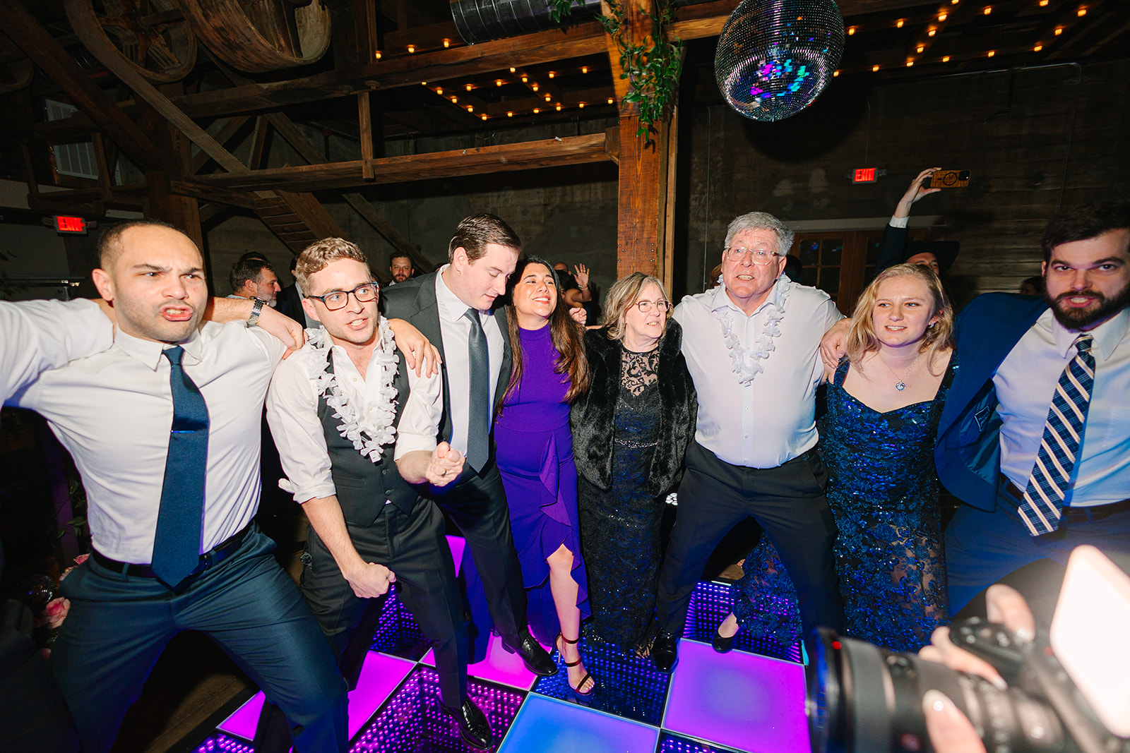 A large group of people dressed formally dance on a colorful light-up floor under disco balls and greenery at a wedding