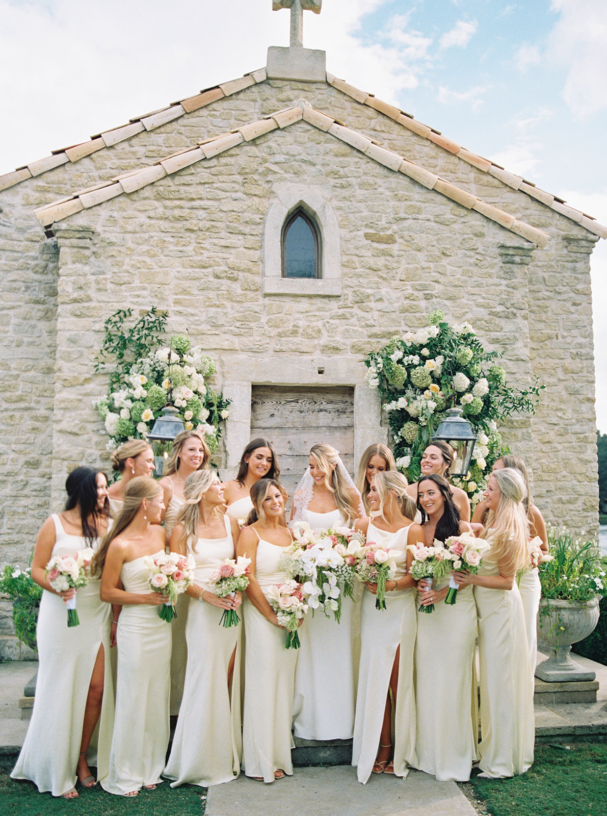 A group of women in matching light-colored dresses stand smiling and holding bouquets in front of a stone building adorned with flowers at The Houston Oaks Country Club