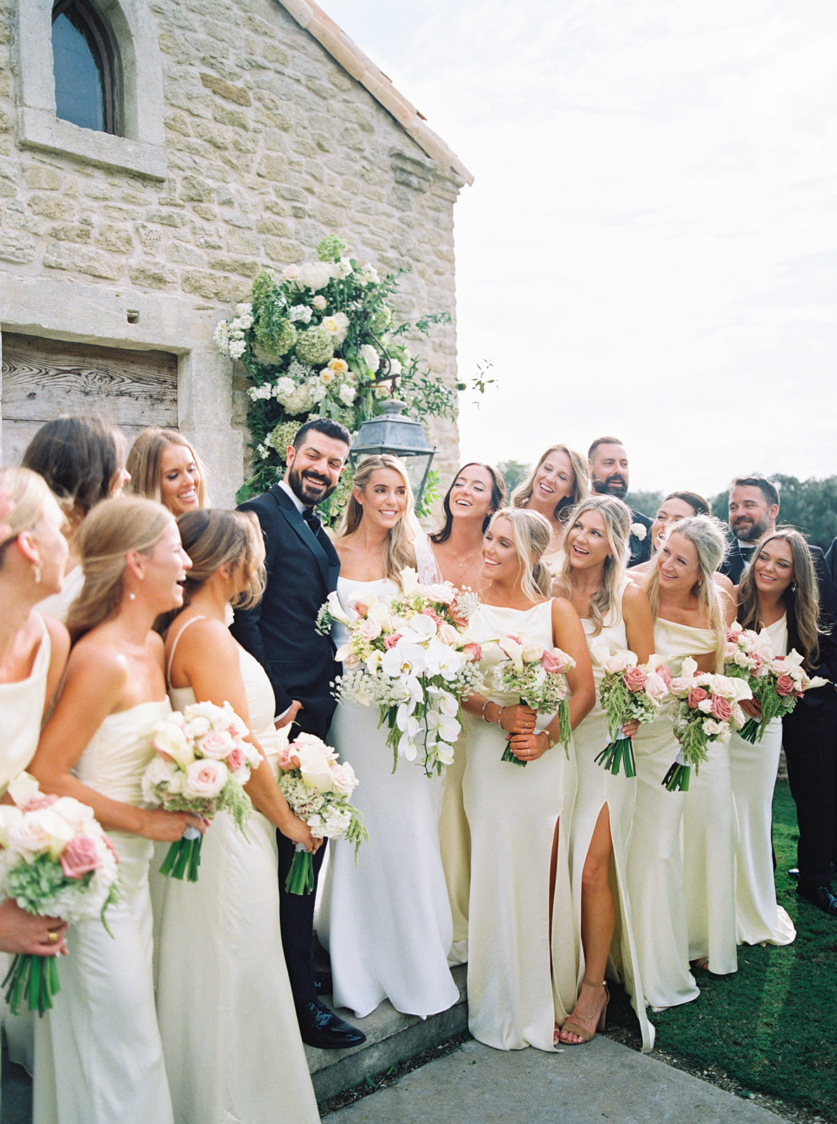 A group of women in matching light-colored dresses stand smiling and holding bouquets in front of a stone building adorned with flowers at The Houston Oaks Country Club