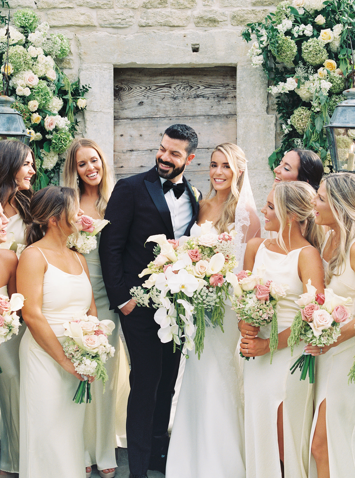 A group of women in matching light-colored dresses stand smiling and holding bouquets in front of a stone building adorned with flowers at The Houston Oaks Country Club