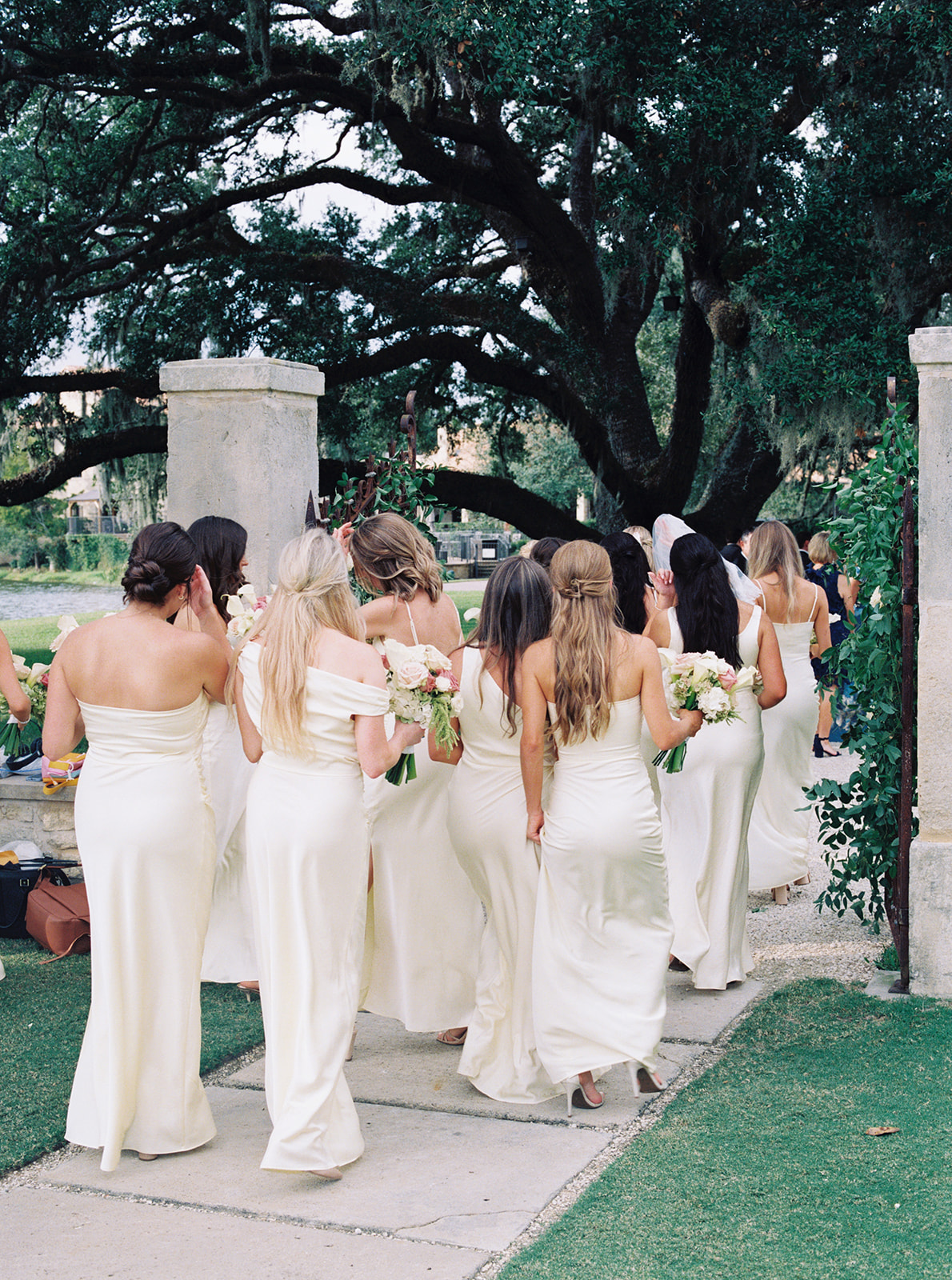 A group of women in matching light-colored dresses stand smiling and holding bouquets in front of a stone building adorned with flowers at The Houston Oaks Country Club