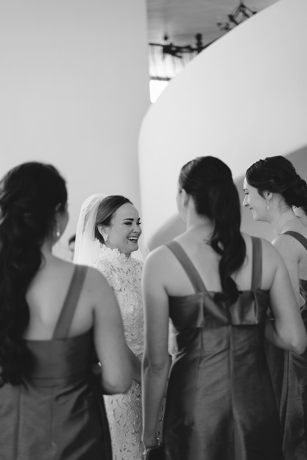 A bride in a white dress stands with eight bridesmaids in green and floral dresses, holding bouquets. They pose indoors against a curved wall and staircase at La Bonne Vie Ranch