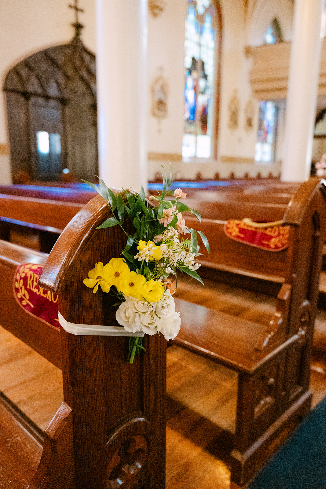 Wooden church pews adorned with small floral arrangements, including yellow and white flowers, in an ornate indoor setting.