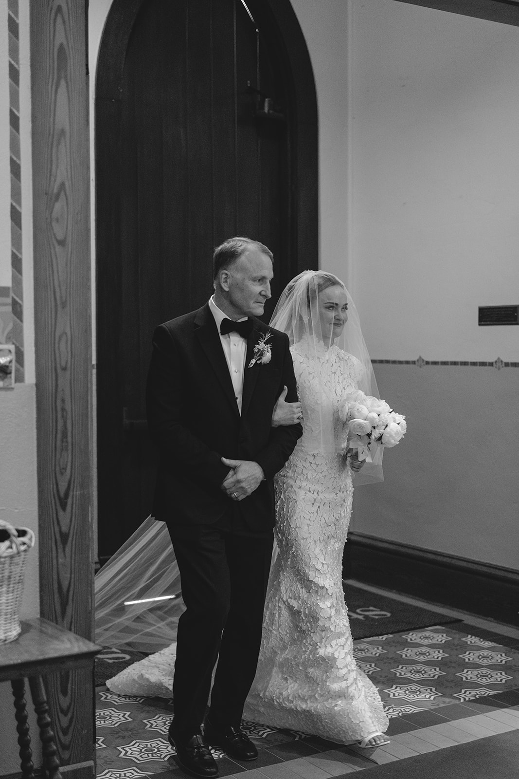A bride in a white gown and veil walks arm in arm with a man in a tuxedo through a doorway.