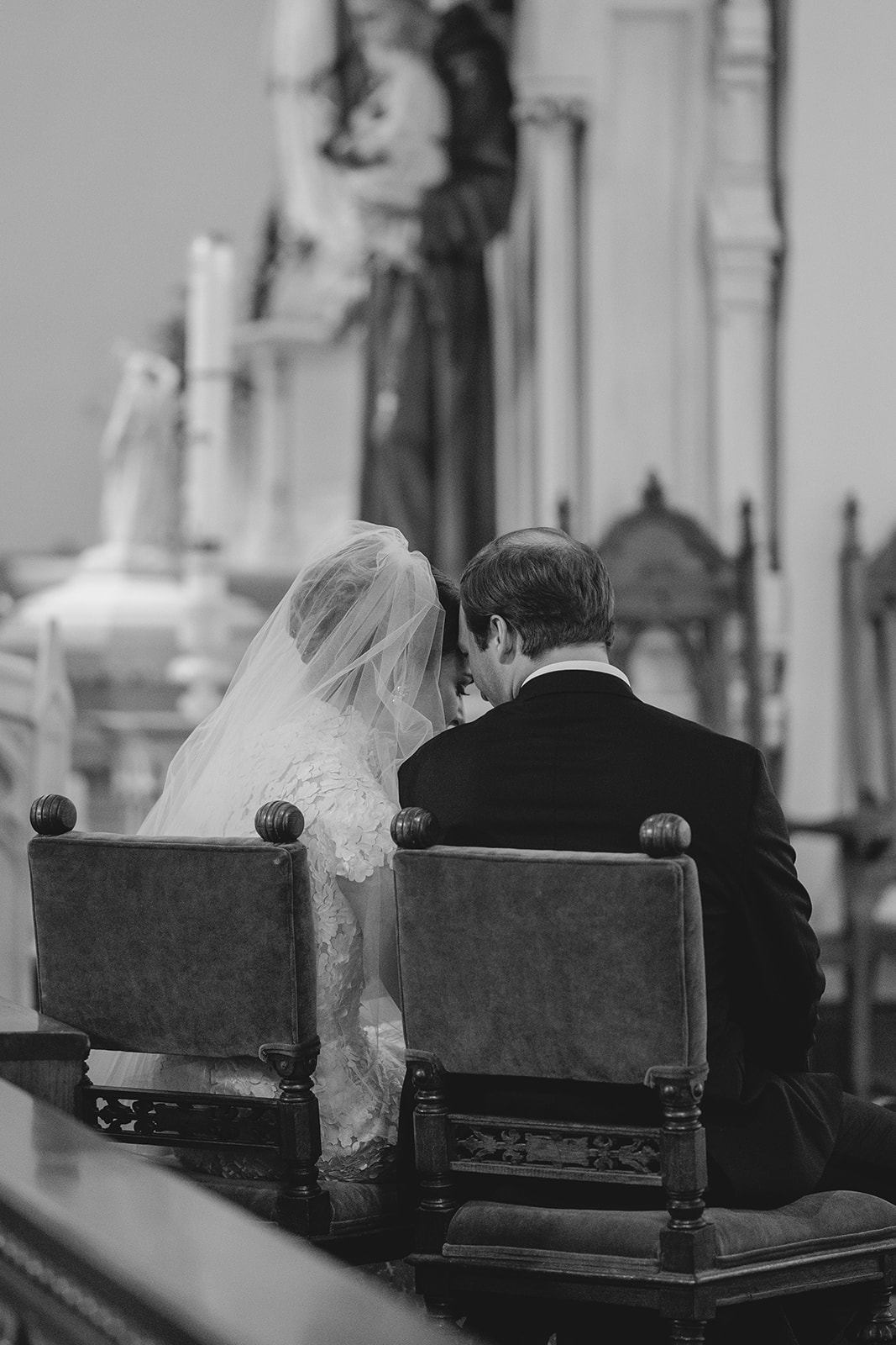 Bride and groom sitting closely on chairs, facing away in a church setting, with statues in the background.