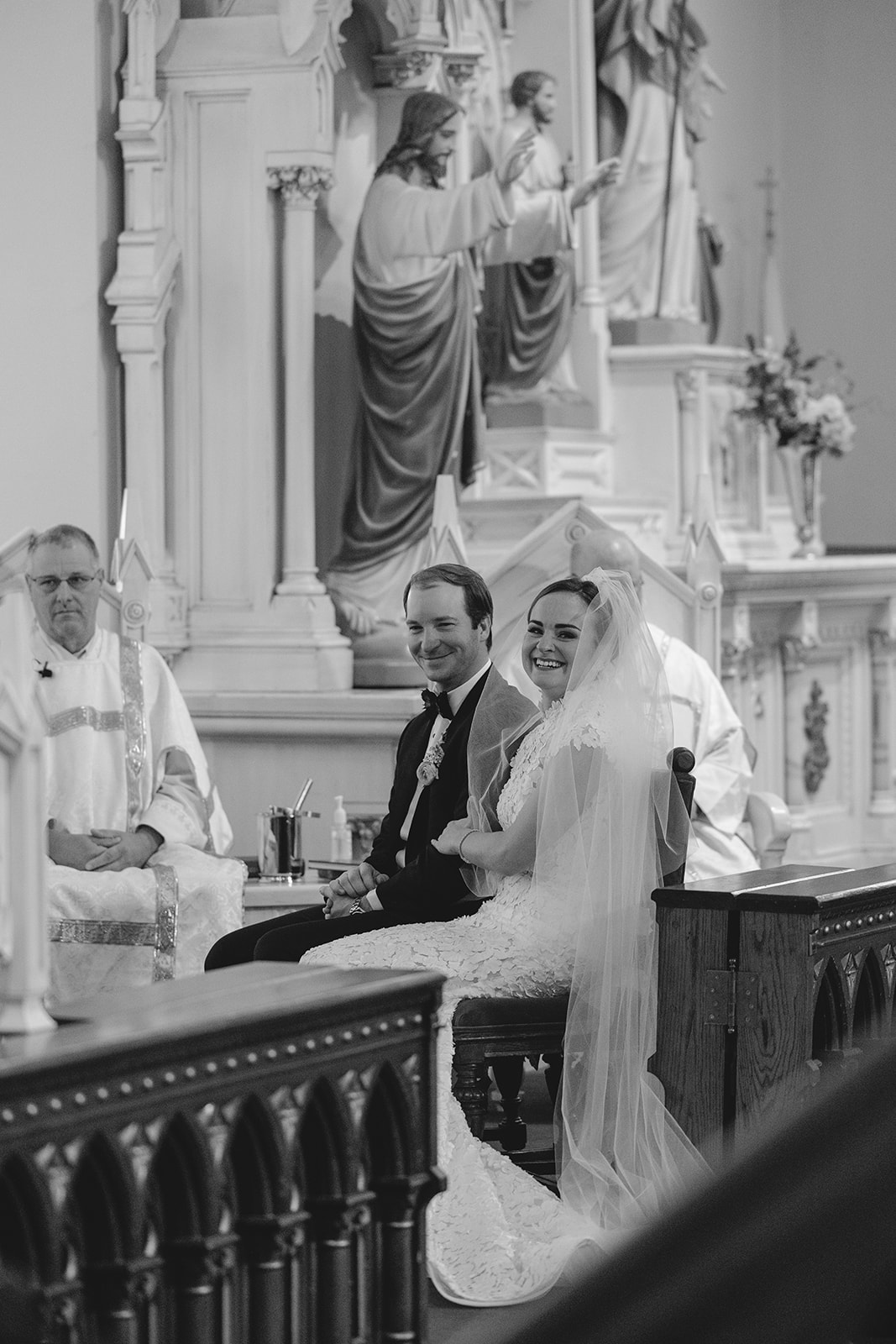 Bride and groom sitting closely on chairs, facing away in a church setting, with statues in the background.