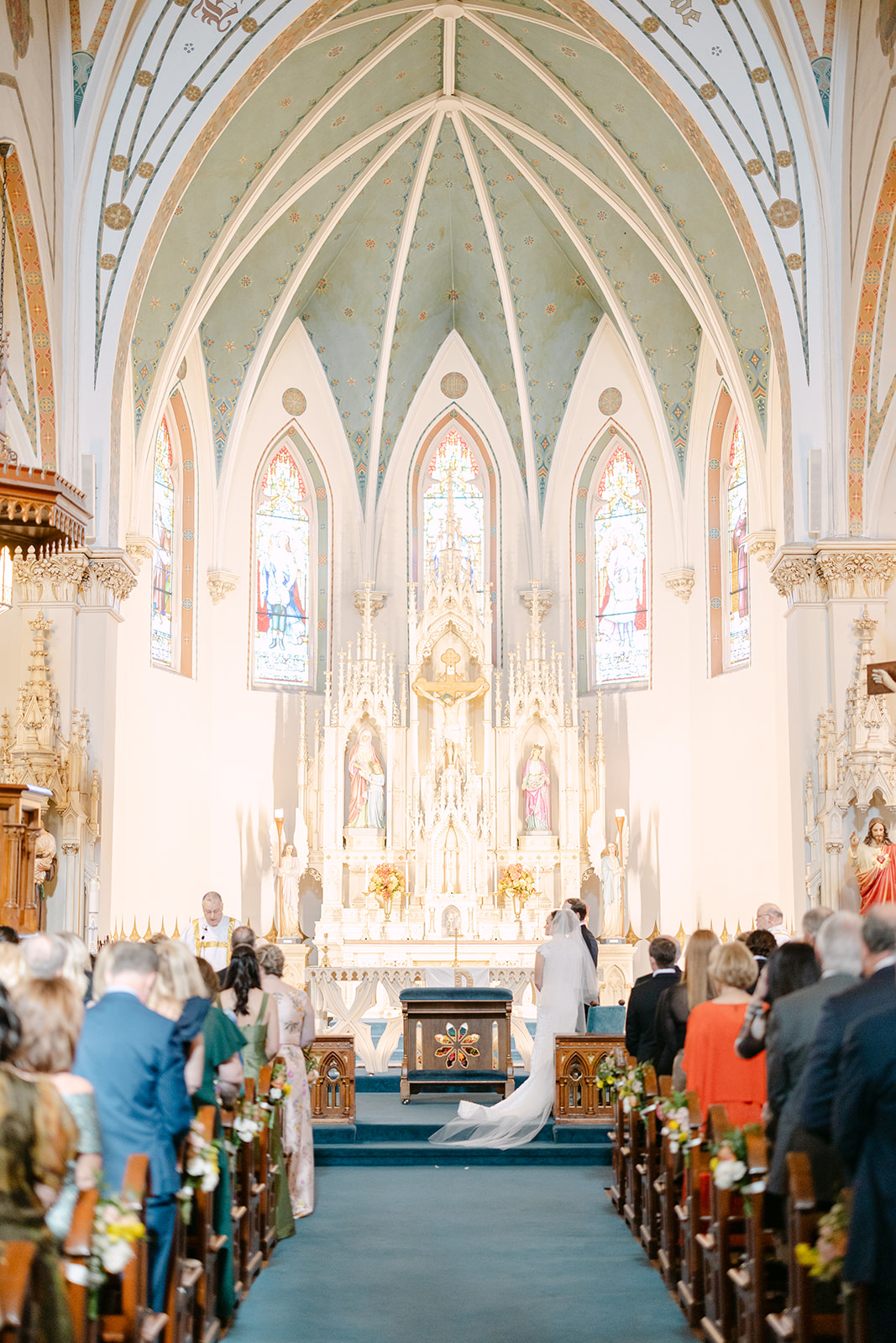 Bride and groom sitting closely on chairs, facing away in a church setting, with statues in the background.