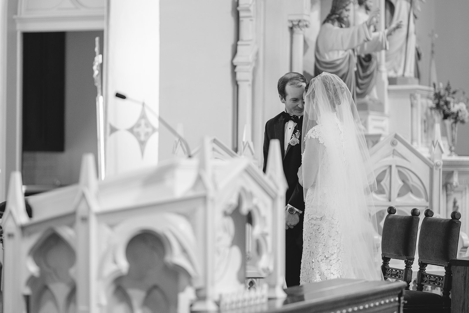Bride and groom sitting closely on chairs, facing away in a church setting, with statues in the background.