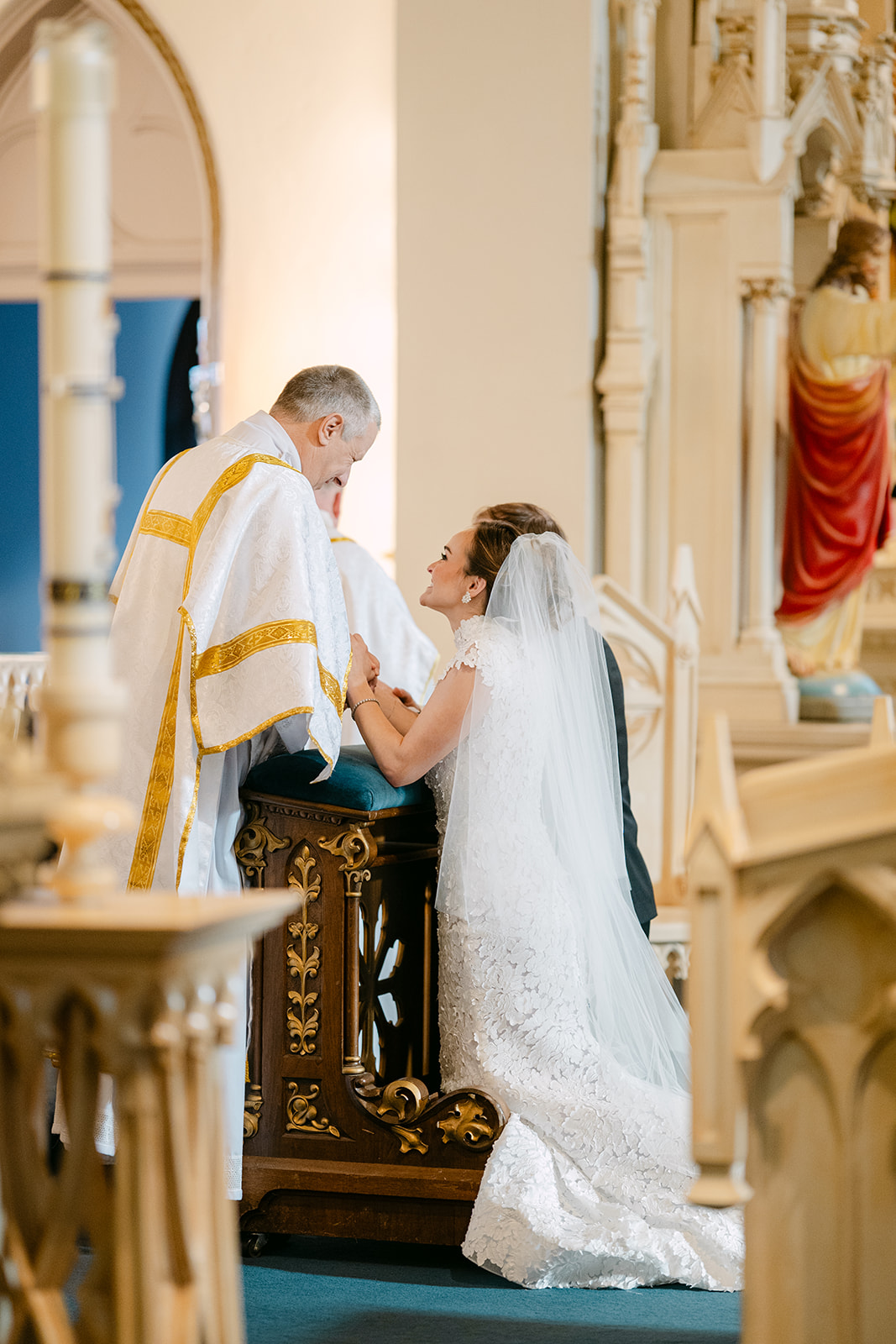 Bride and groom sitting closely on chairs, facing away in a church setting, with statues in the background.