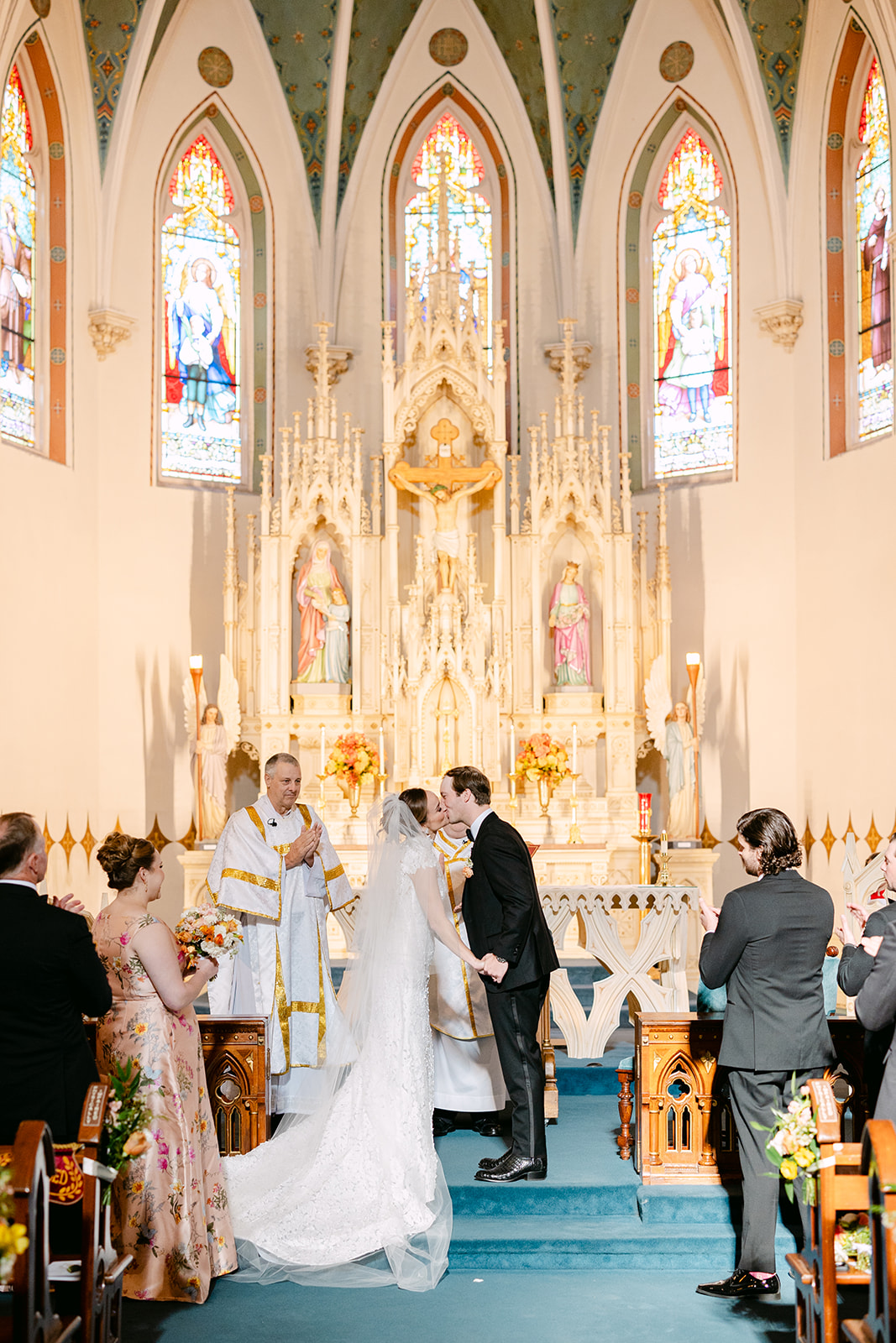 Bride and groom sitting closely on chairs, facing away in a church setting, with statues in the background.