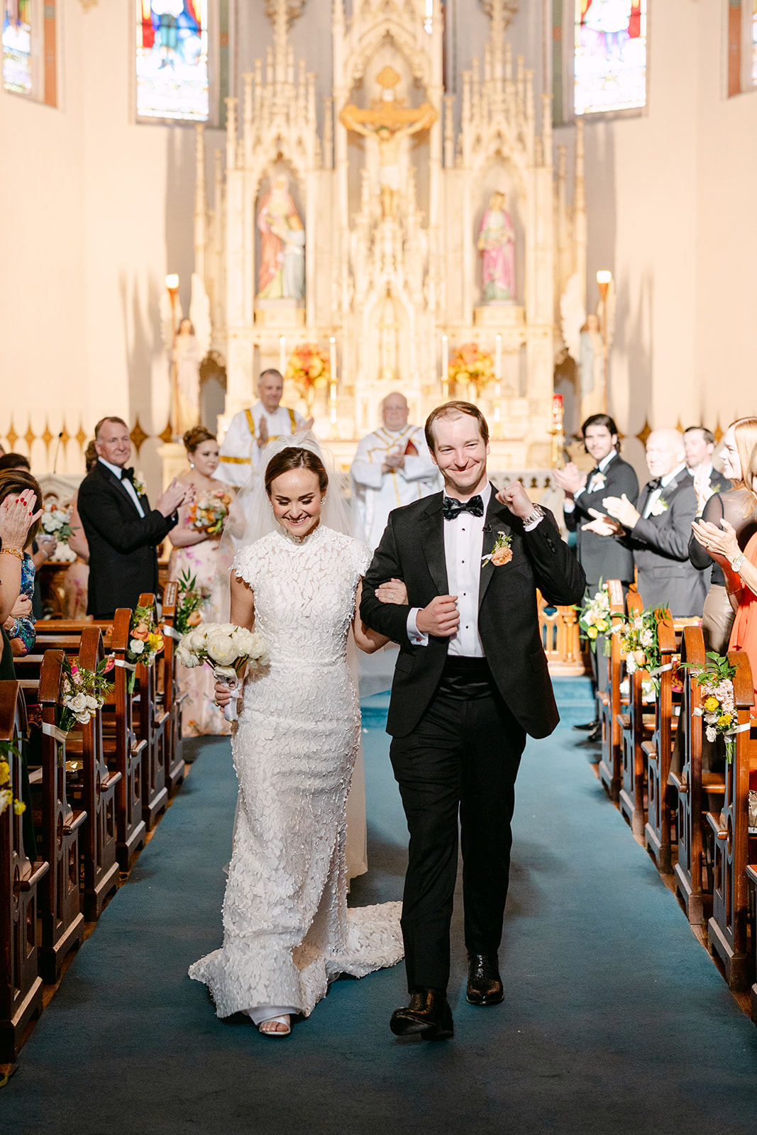 Bride and groom walk down the aisle in a church, smiling and holding hands, surrounded by clapping guests.