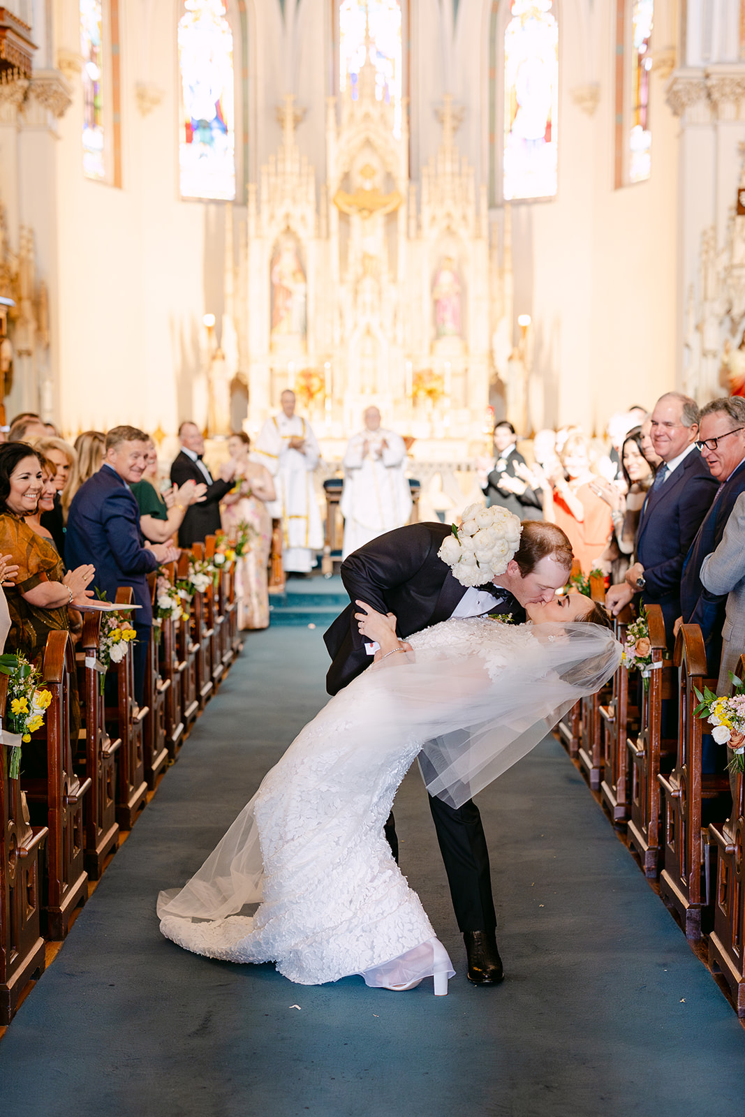 Bride and groom walk down the aisle in a church, smiling and holding hands, surrounded by clapping guests.