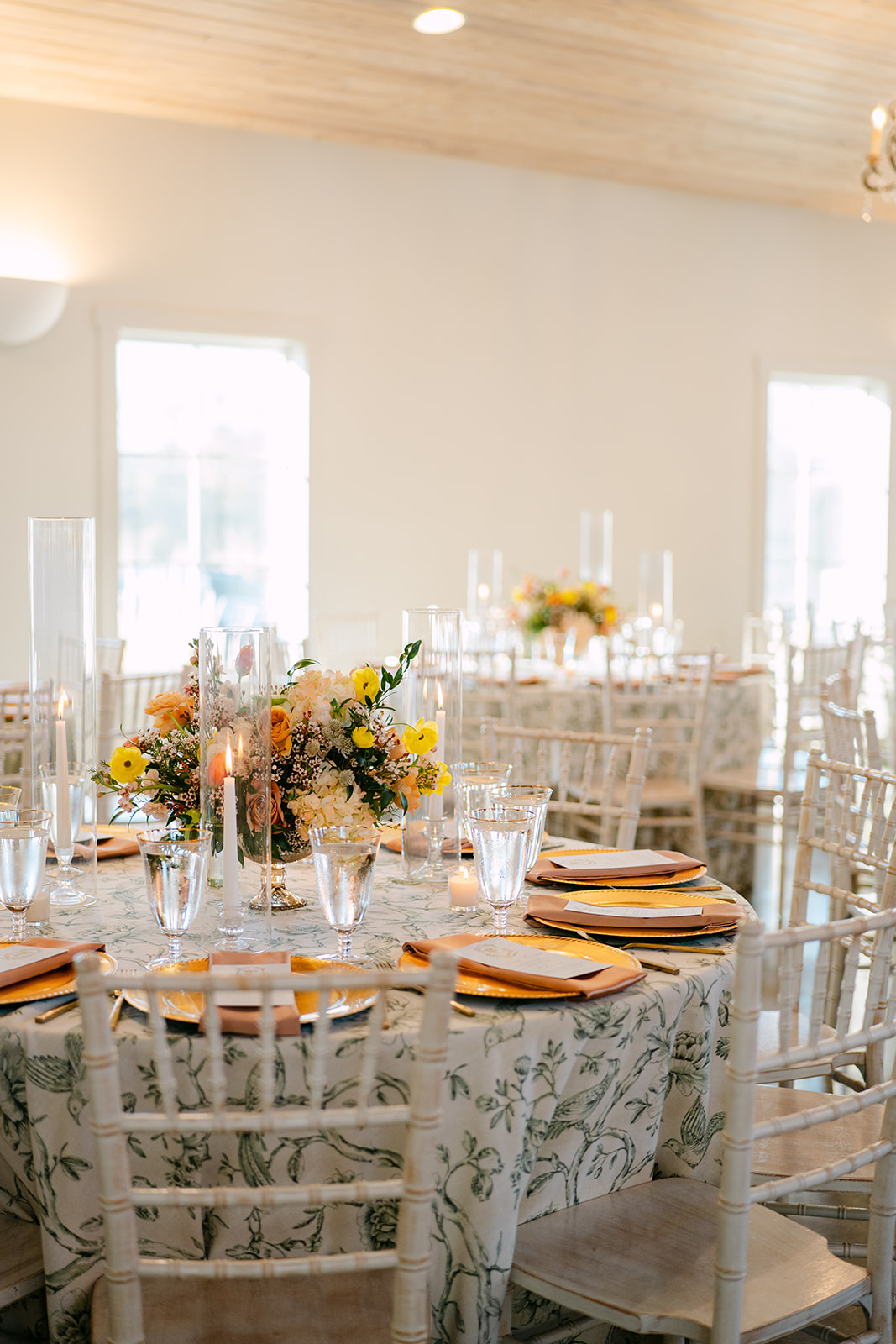 Elegant table setting with a floral centerpiece, surrounded by candles, glasses, and place settings with menus on gold chargers. White chairs encircle the table.