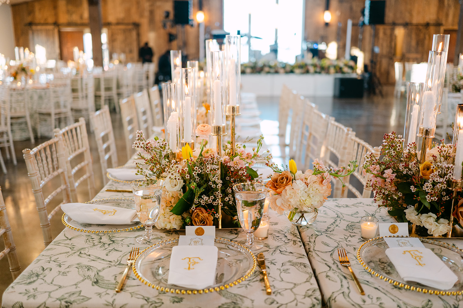 Elegant table setting with a floral centerpiece, surrounded by candles, glasses, and place settings with menus on gold chargers. White chairs encircle the table.