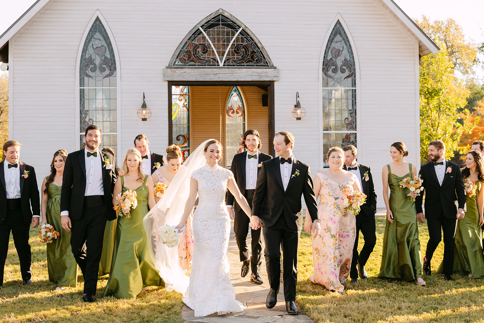 A wedding party stands outside a church. The bride and groom hold hands, surrounded by bridesmaids in green dresses and groomsmen in black suits at La Bonne Vie Ranch