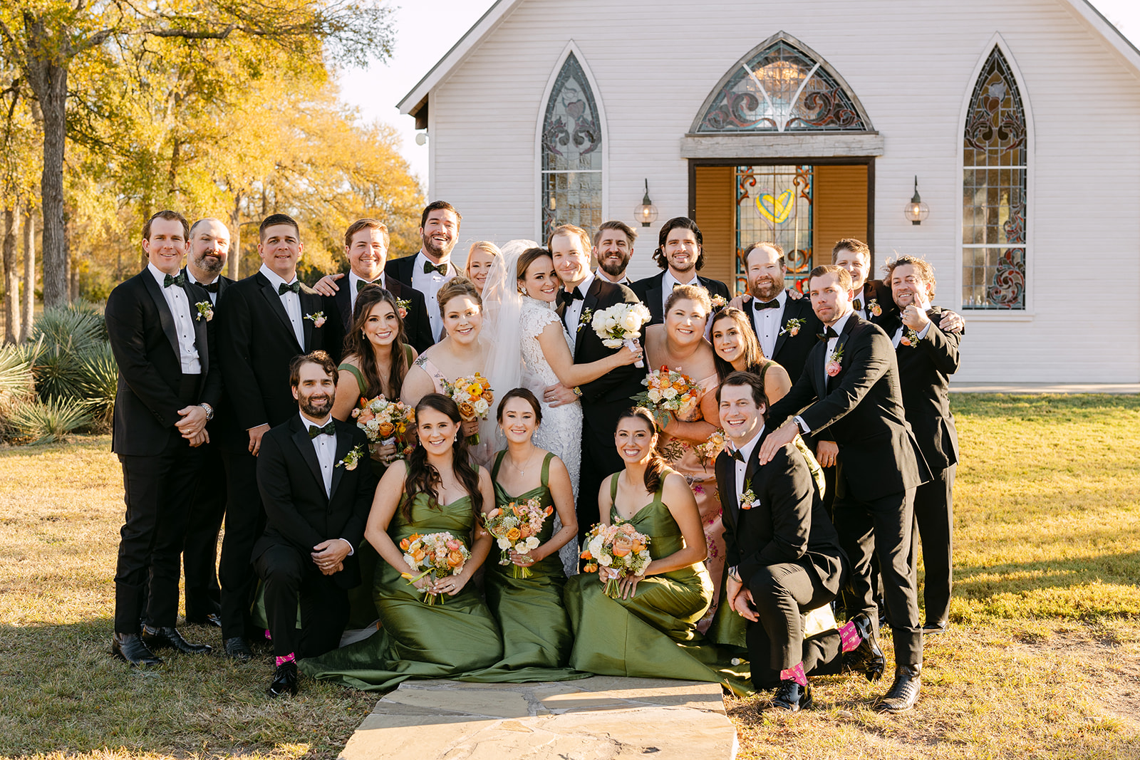 A wedding party stands outside a church. The bride and groom hold hands, surrounded by bridesmaids in green dresses and groomsmen in black suits at La Bonne Vie Ranch