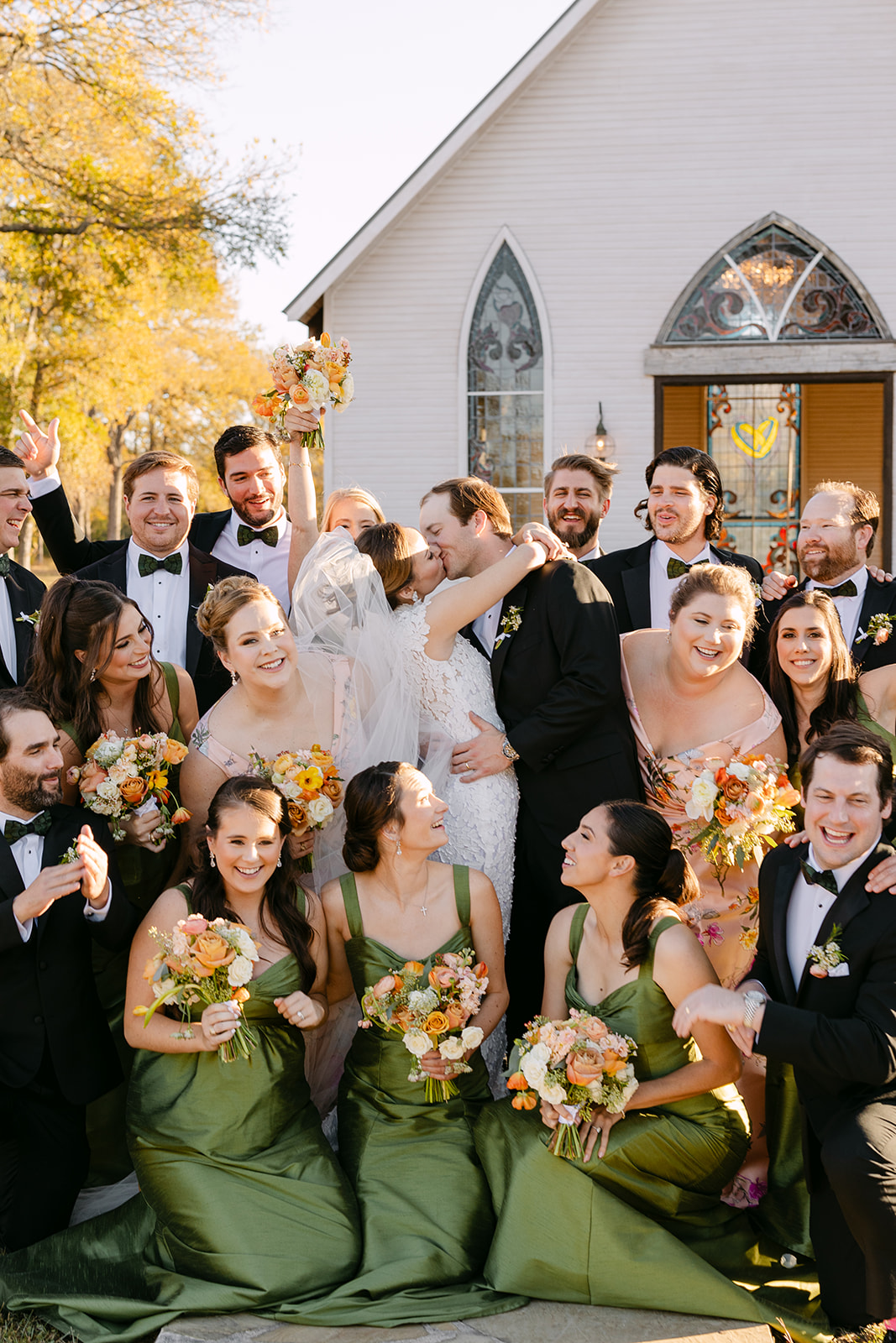A wedding party stands outside a church. The bride and groom hold hands, surrounded by bridesmaids in green dresses and groomsmen in black suits at La Bonne Vie Ranch