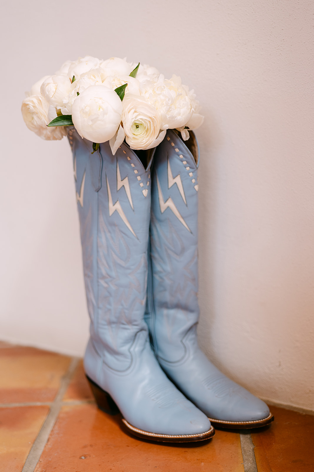 Blue cowboy boots with white lightning bolt designs hold a bouquet of white flowers at the top, placed on a tile floor.