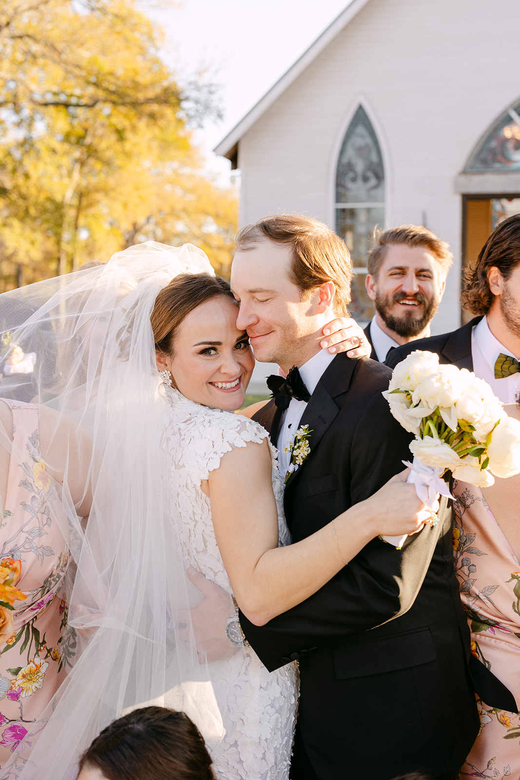 A wedding party stands outside a church. The bride and groom hold hands, surrounded by bridesmaids in green dresses and groomsmen in black suits at La Bonne Vie Ranch