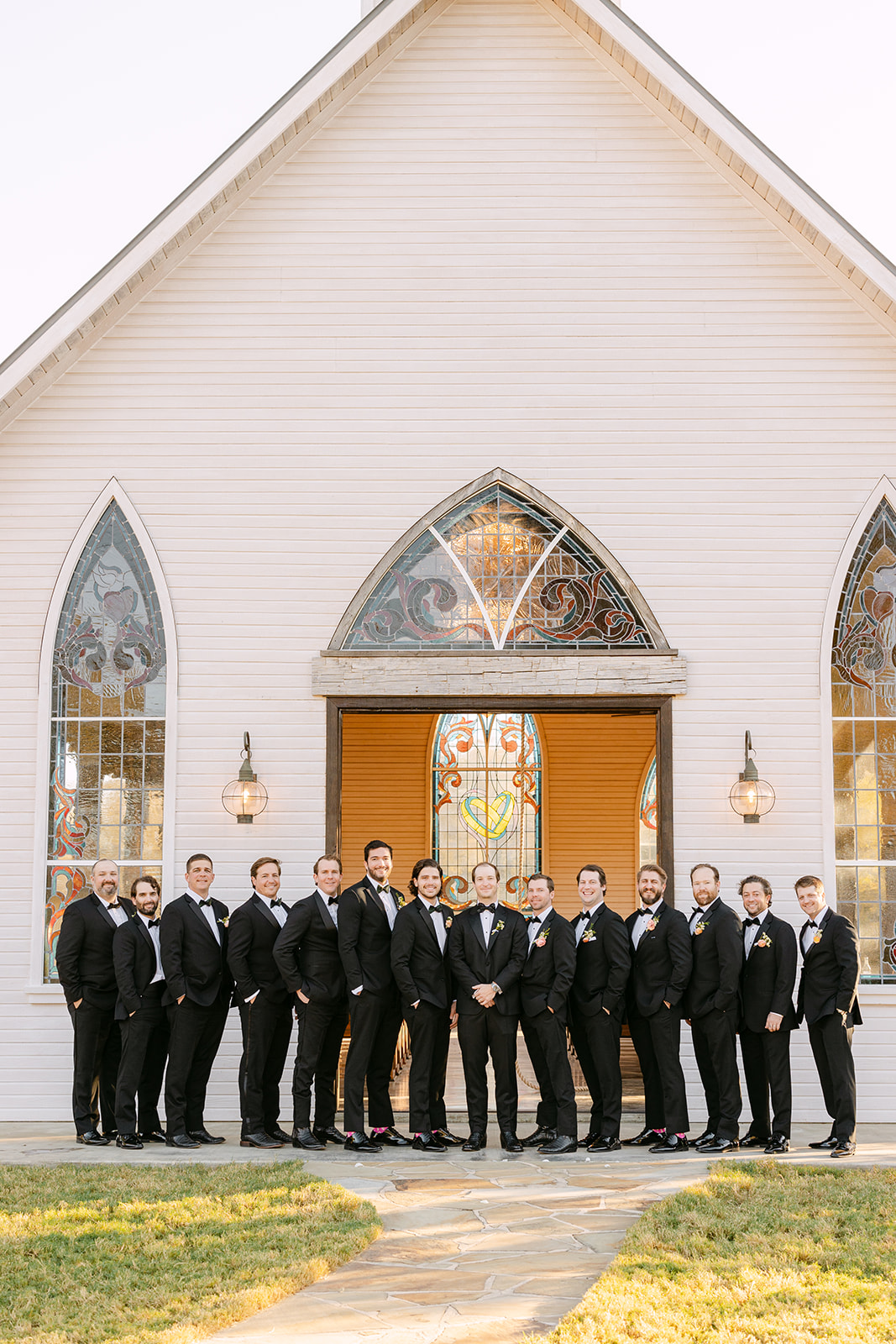 A group of men in formal suits stand in a line outside a building with arched stained glass windows and wooden double doors.