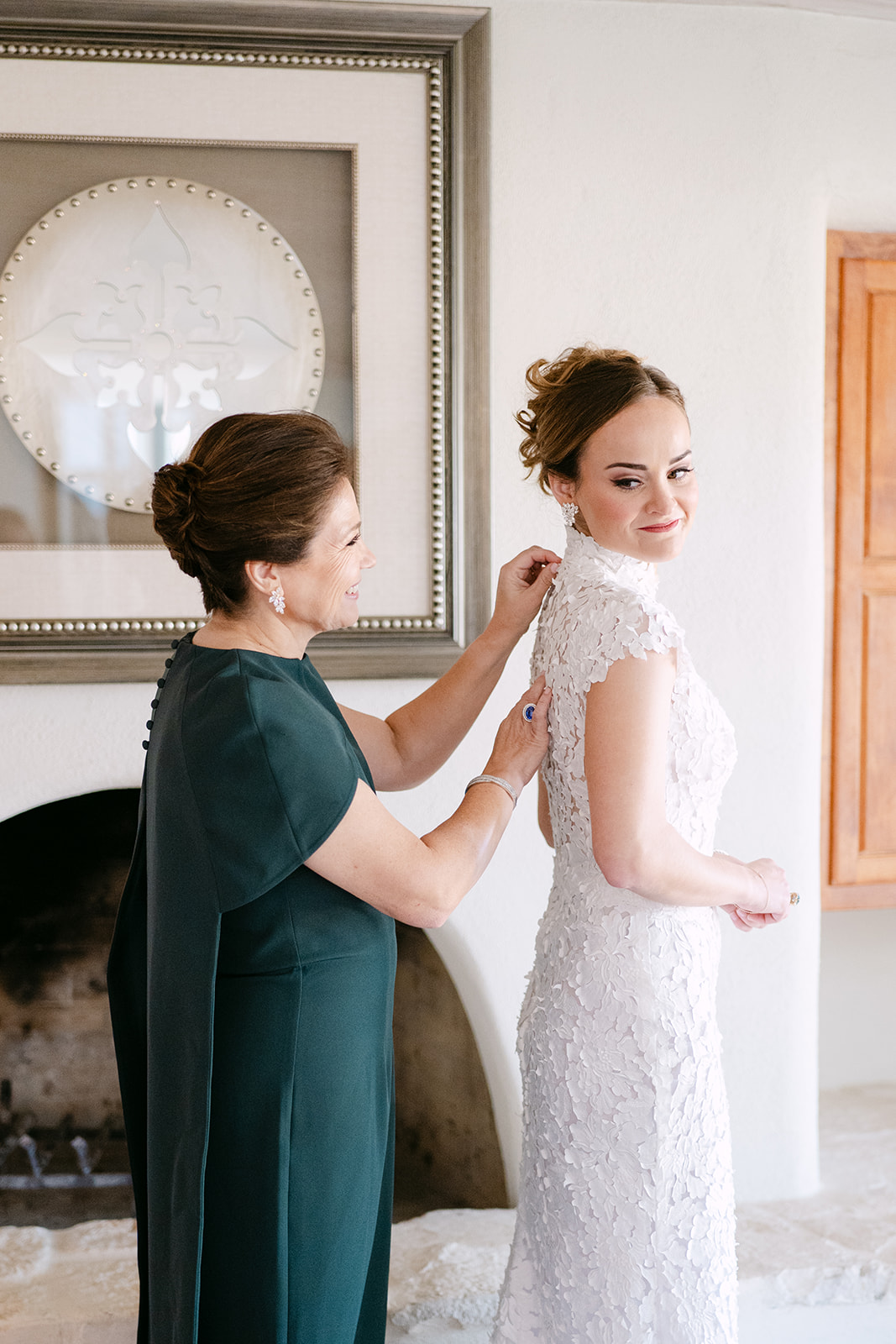 A woman in a dark dress helps a bride in a lace gown with her dress. The bride smiles at the camera, standing in a room with a framed decorative plate on the wall.