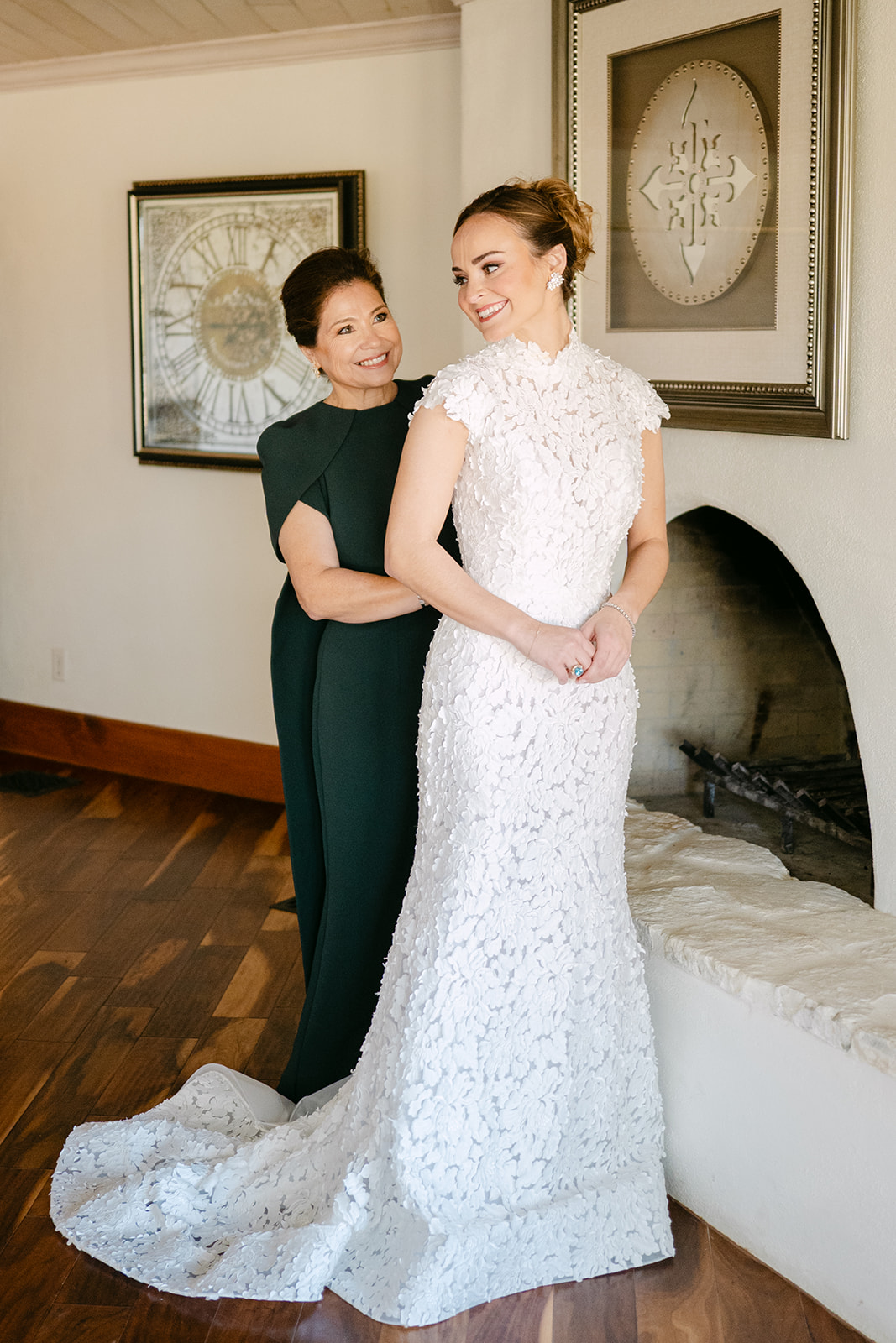 A woman in a dark dress helps a bride in a lace gown with her dress. The bride smiles at the camera, standing in a room with a framed decorative plate on the wall.