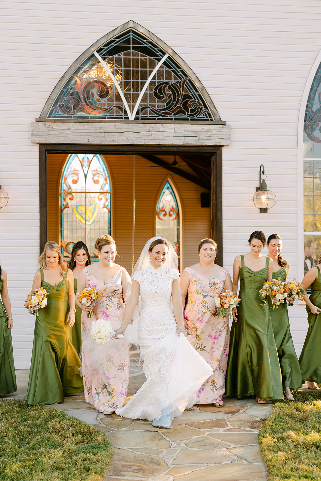 A bride in a white dress stands with eight bridesmaids in green and floral dresses, holding bouquets. They pose indoors against a curved wall and staircase at La Bonne Vie Ranch
