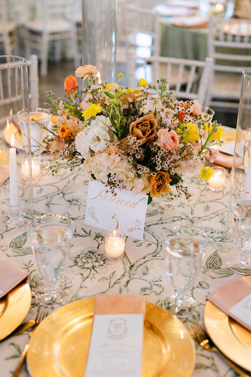 Elegant table setting with a floral centerpiece, surrounded by candles, glasses, and place settings with menus on gold chargers. White chairs encircle the table.